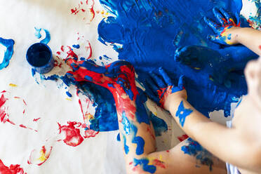 Toddler girl sitting on paper on the floor playing with finger paint, partial view - GEMF03024