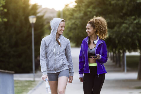 Zwei lächelnde sportliche junge Frauen gehen nach dem Training im Park spazieren, lizenzfreies Stockfoto