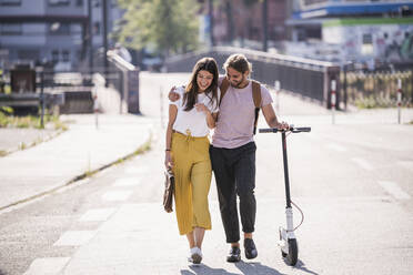 Happy young couple with electric scooter walking on the street - UUF18398