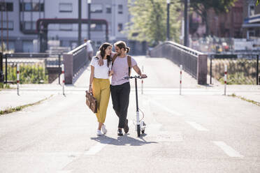 Glückliches junges Paar mit Elektroroller zu Fuß auf der Straße - UUF18399