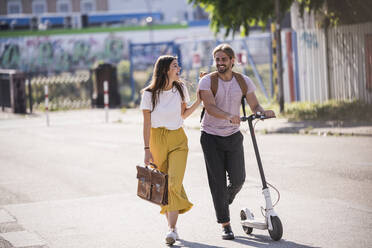 Glückliches junges Paar mit Elektroroller zu Fuß auf der Straße - UUF18400