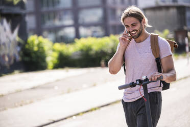 Happy young man with electric scooter talking on the phone on the street - UUF18402