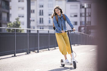 Glückliche junge Frau auf einem Elektroroller auf einer Brücke - UUF18411