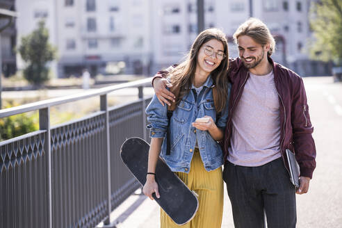 Glückliches junges Paar mit Skateboard und Smartphone auf einer Brücke - UUF18415