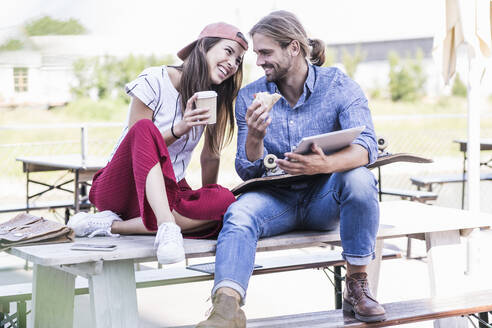 Junges Paar sitzt an einem Tisch in einem Biergarten mit Tablet, Sandwich und Kaffee zum Mitnehmen - UUF18438