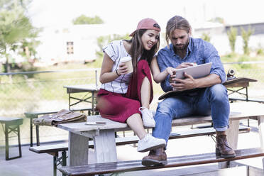 Young couple sitting on table in a beer garden using tablet - UUF18439