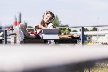 Young woman relaxing in a beer garden with headphones and tablet - UUF18450