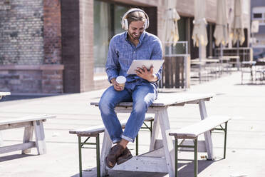 Junger Mann sitzt mit Kopfhörern und Tablet auf einem Tisch in einem Biergarten - UUF18453