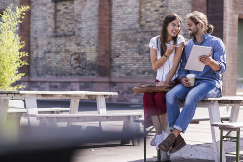 Glückliches junges Paar sitzt am Tisch in einem Biergarten mit Handy und Tablet, lizenzfreies Stockfoto