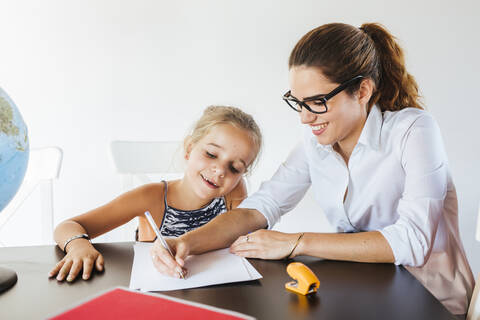 Lehrerin hilft Schülerin am Schreibtisch beim Schreiben auf Papier, lizenzfreies Stockfoto