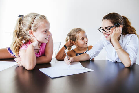 Glückliche Lehrerin am Schreibtisch sitzend mit zwei Schulmädchen, die auf Papier schreiben, lizenzfreies Stockfoto