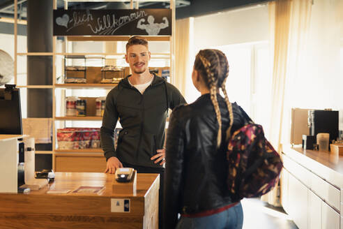 Trainer im Gespräch mit junger Frau an der Rezeption eines Fitnessstudios - SEBF00119