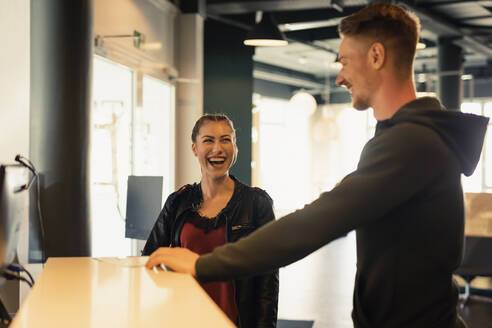 Lachende junge Frau im Gespräch mit dem Trainer an der Rezeption eines Fitnessstudios - SEBF00121