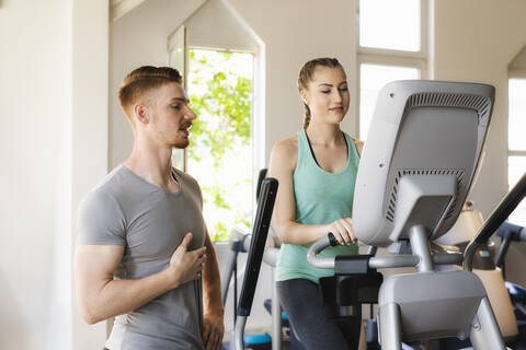 Personal Trainer im Gespräch mit einer jungen Frau, die auf einem Step-Gerät im Fitnessstudio trainiert, lizenzfreies Stockfoto