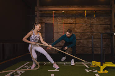 Young woman exercising in fitness gym with personal trainer pulling rope - SEBF00140