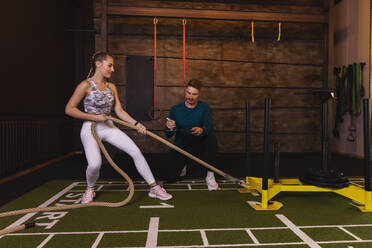 Junge Frau trainiert in einem Fitnessstudio mit einem persönlichen Trainer, der ein Seil zieht - SEBF00142