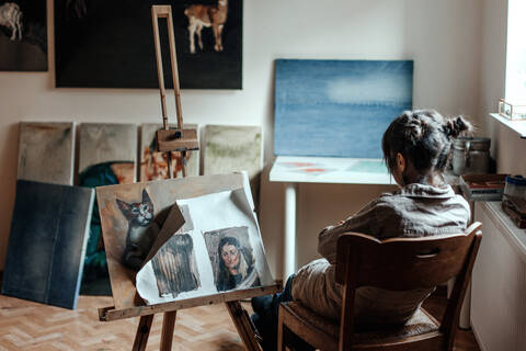 Rückansicht einer Künstlerin, die in ihrem Atelier vor einem Gemälde sitzt, lizenzfreies Stockfoto