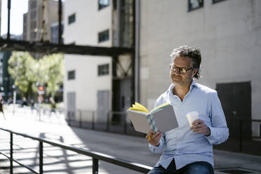 Porträt eines reifen Mannes, der ein Buch liest und eine Tasse Kaffee zum Mitnehmen hält - KNSF06133
