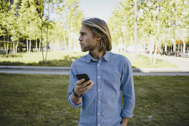 Junger Mann mit Handy in einem Park, der sich umschaut - KNSF06161