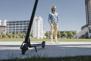 Smiling young man standing next to e-scooter in the city - KNSF06170