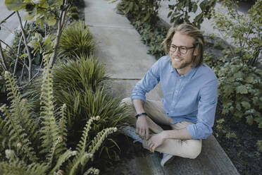 Lächelnder junger Mann sitzt auf dem Bürgersteig, umgeben von Pflanzen - KNSF06172