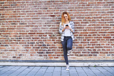 Young woman leaning against brick wall in the city while using smartphone - BSZF01316