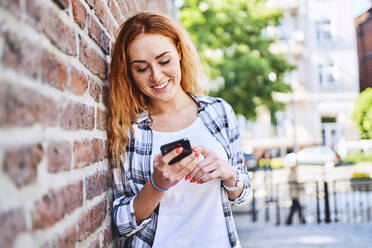 Junge Frau lehnt sich in der Stadt an eine Mauer, während sie ihr Smartphone benutzt - BSZF01317