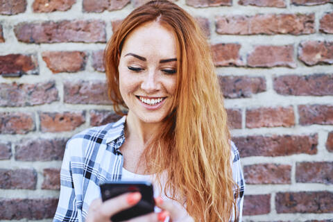 Junge Frau lehnt sich in der Stadt an eine Mauer, während sie ihr Smartphone benutzt, lizenzfreies Stockfoto