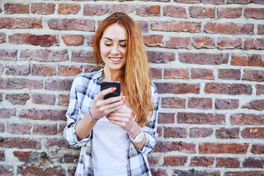 Cheerful young woman using smartphone while leaning against brick wall - BSZF01319