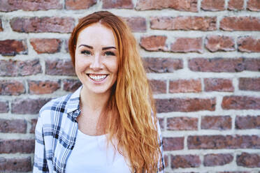 Portrait of beautiful young woman smiling at camera in front of brick wall - BSZF01320