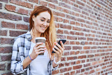 Young woman standing against brick wall using smartphone and holding coffee cup - BSZF01321