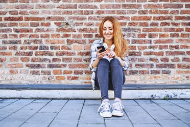 Lächelnde junge Frau, die in der Stadt im Freien sitzt und ihr Smartphone benutzt - BSZF01326