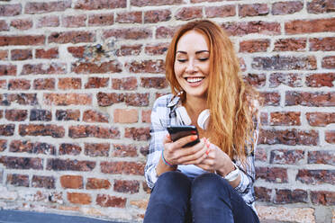 Fröhliche junge Frau, die an einer Backsteinmauer sitzt und ihr Smartphone benutzt - BSZF01329