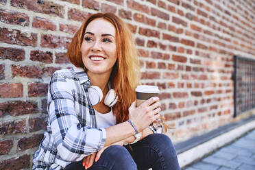 Junge Frau sitzt an einer Backsteinmauer, hält eine Tasse Kaffee und schaut weg - BSZF01330