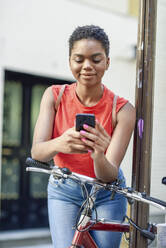 Portrait of smiling young woman leaning on handle bar of bicycle looking at cell phone - JSMF01206