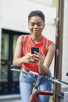 Porträt einer lächelnden jungen Frau, die sich auf den Lenker eines Fahrrads stützt und auf ihr Mobiltelefon schaut - JSMF01206