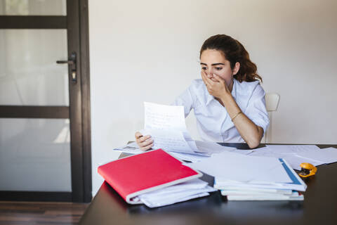 Schockierte Studentin beim Lesen eines Dokuments am Schreibtisch zu Hause, lizenzfreies Stockfoto