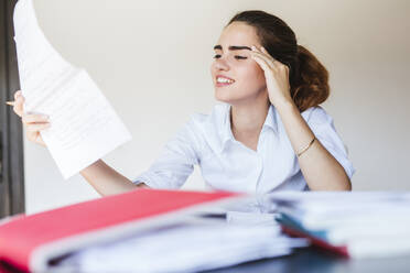 Smiling female student reading document at desk at home - LJF00601