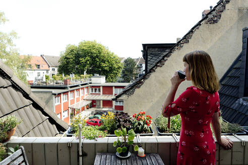Junge Frau entspannt sich mit einer Tasse Kaffee auf dem Balkon und schaut in die Ferne - FLLF00259