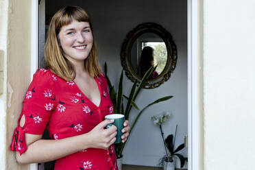 Porträt einer lächelnden jungen Frau mit einer Tasse Kaffee auf einem Balkon stehend - FLLF00260
