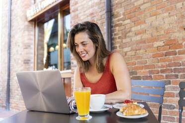 Lächelnde junge Bloggerin sitzt im Straßencafé und arbeitet am Laptop - AFVF03633