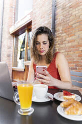Porträt eines jungen Bloggers, der in einem Straßencafé sitzt und auf sein Handy schaut - AFVF03635