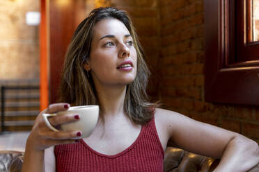 Porträt einer jungen Frau mit einer Tasse Kaffee, die aus dem Fenster schaut - AFVF03648