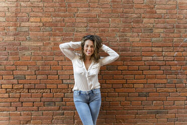 Porträt einer lächelnden Frau vor einer Backsteinmauer, die Hände im Haar - AFVF03652