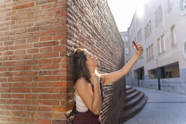 Junge Frau lehnt an einer Backsteinmauer und macht ein Selfie - AFVF03707