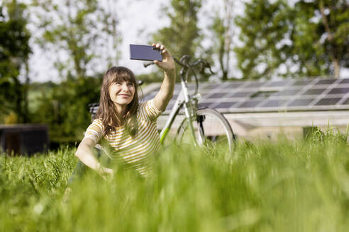 Entspannte Frau sitzt auf einer Wiese mit Fahrrad und macht ein Selfie - FMKF05773