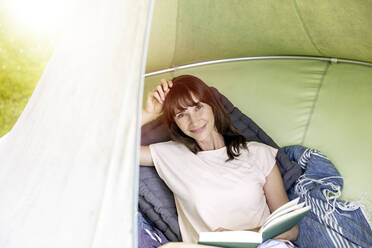 Portrait of smiling woman reading book in a hanging tent - FMKF05793