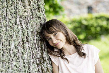 Portrait of smiling brunette woman leaning against a tree trunk - FMKF05799