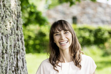 Portrait of happy brunette woman at a tree trunk - FMKF05803