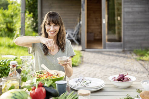 Portrait of smiling woman preparing a salad on garden table - FMKF05804